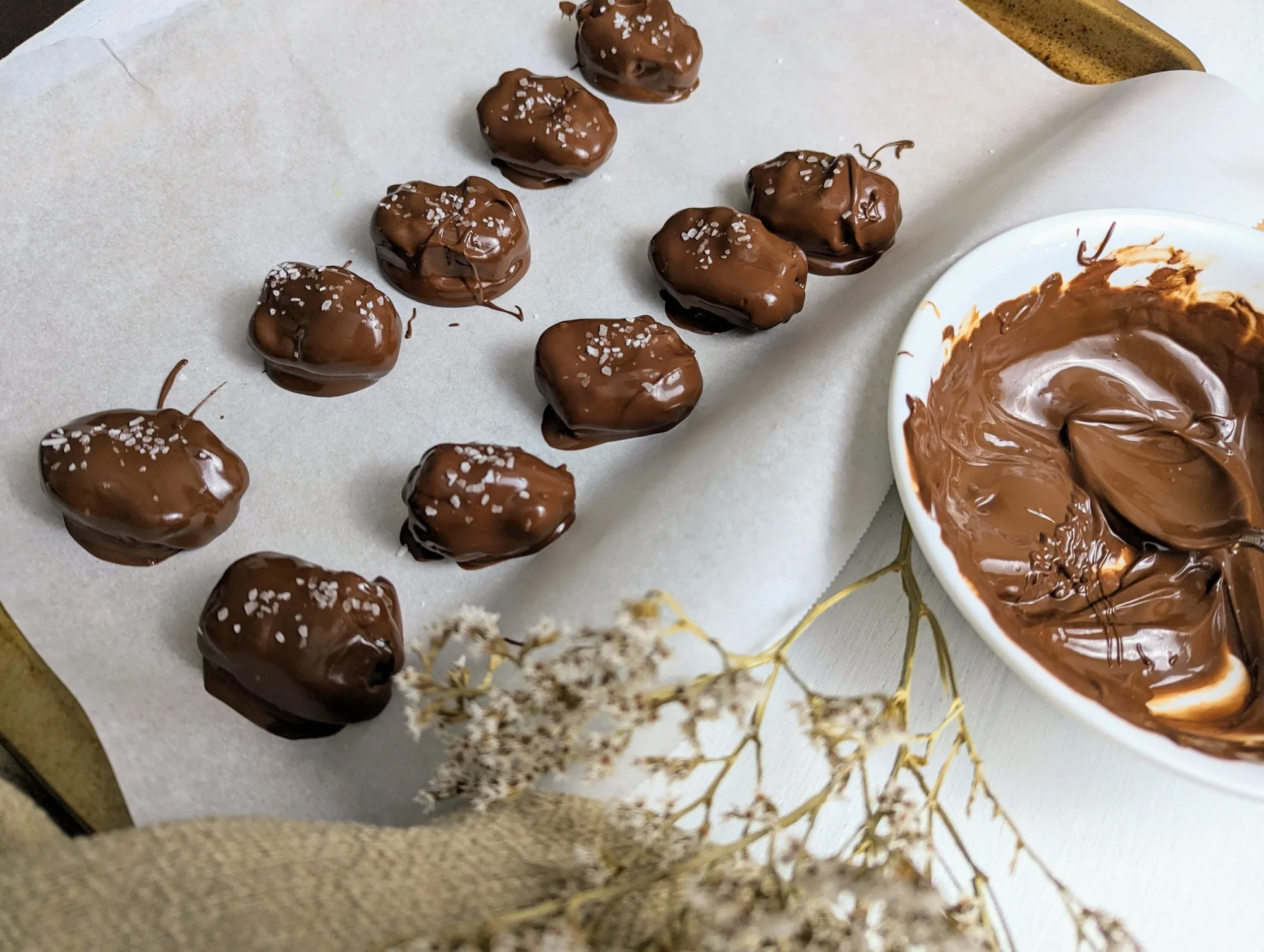 Chocolate covered dates on a parchment-lined baking sheet next to a bowl of melted chocolate