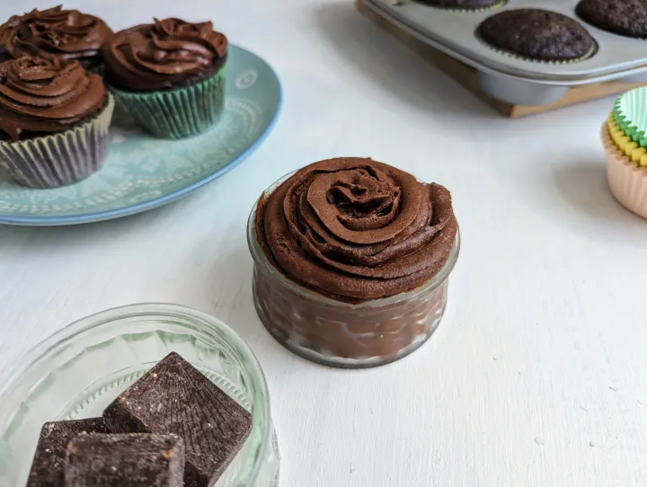 A glass jar filled with a swirl of chocolate frosting, next to some cupcakes and a few pieces of chocolate