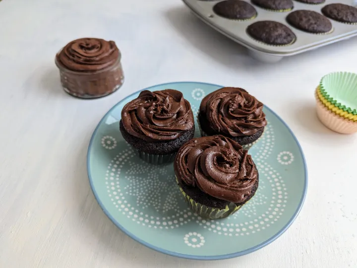Chocolate cupcakes with chocolate frosting on a blue plate, next to a jar of frosting and a cupcake tin full of cupcakes.