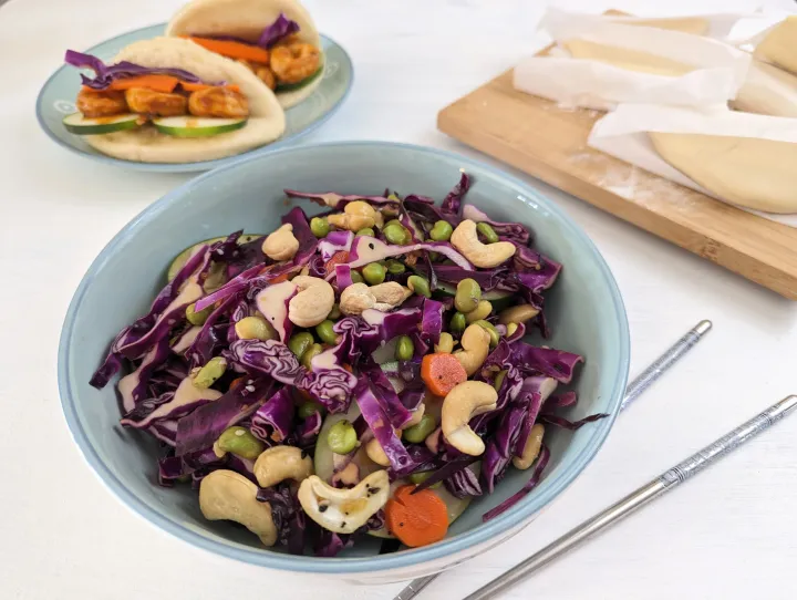 A red cabbage salad with cashews, edamame, carrot and cucumber in a blue bowl, with bao buns in the background