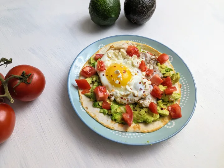 Toasted tortilla on a plate with smashed avocado, chopped tomato, a fried egg and fried feta