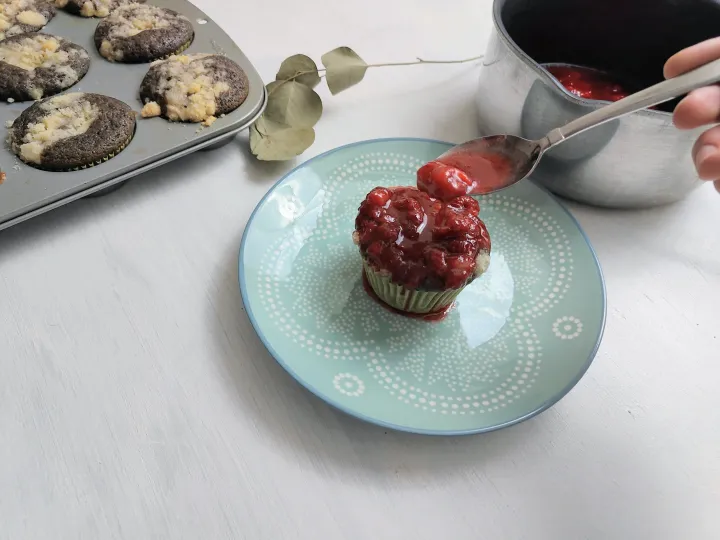 A strawberry topped muffin on a plate next to a saucepan of strawberry syrup and a muffin tin of muffins.